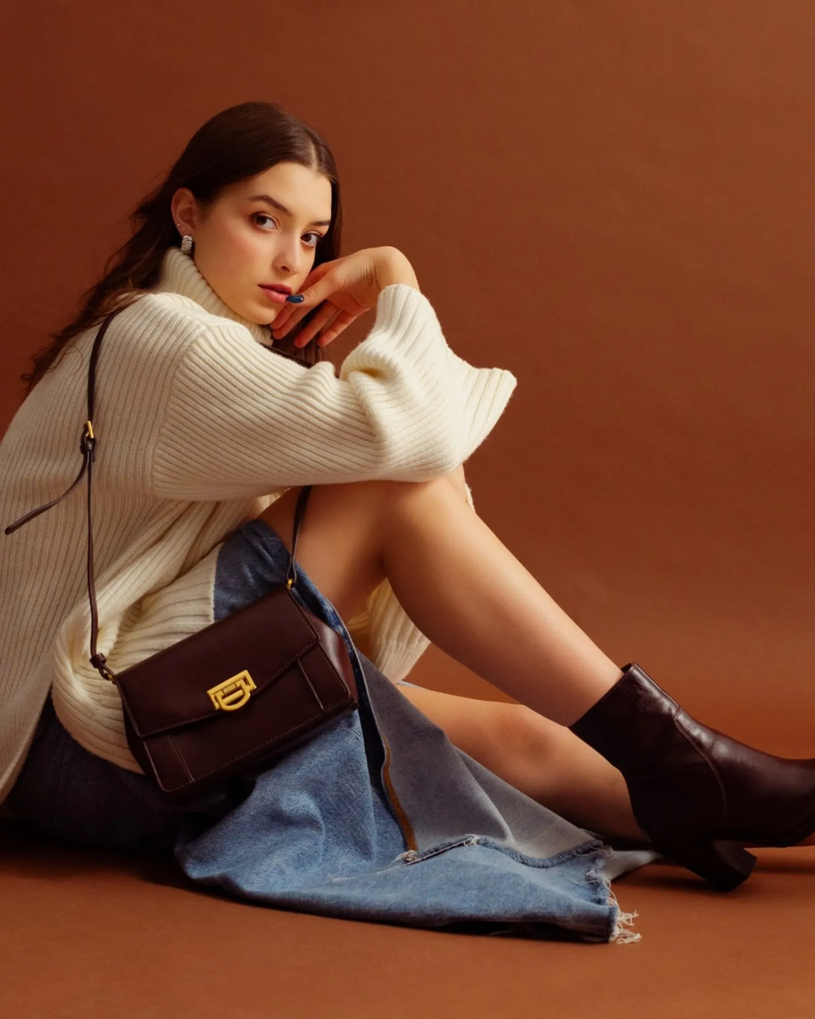 A young woman in a cream sweater and denim sitting against a warm rust-colored studio backdrop